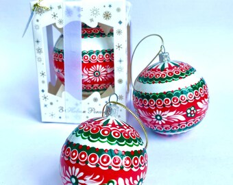 May include: Two hand-painted Christmas baubles with red, white and green designs. The baubles are round and feature a traditional folk art style. The baubles are in a white box with a gold snowflake design.