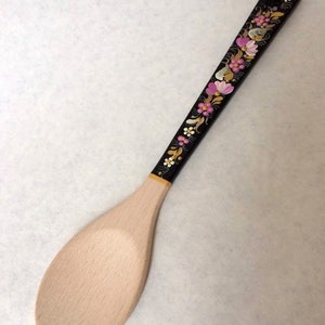 May include: Wooden spoon with a light-colored, oval-shaped bowl and a long, black handle. The handle is decorated with a floral pattern in pink, white, and yellow. The spoon is likely used for cooking and serving.