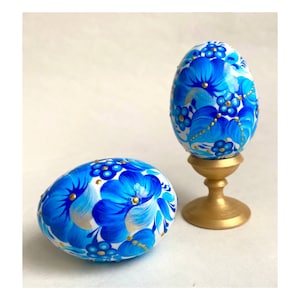 Puede incluir: Dos huevos de Pascua de madera pintados a mano con diseños florales azules y dorados. El huevo de la izquierda está sobre una superficie blanca, mientras que el huevo de la derecha está sobre un soporte dorado.