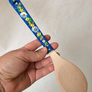 May include: A wooden spatula with a blue handle decorated with colorful floral designs. The spatula's handle is painted blue and adorned with white, yellow, and blue flowers. The spatula's blade is made of light-colored wood.