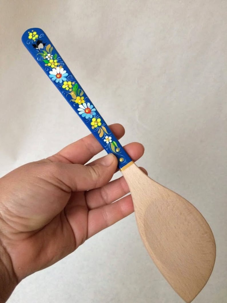 May include: A wooden spatula with a blue handle decorated with colorful floral designs. The spatula's handle is painted blue and adorned with white, yellow, and blue flowers. The spatula's blade is made of light-colored wood.