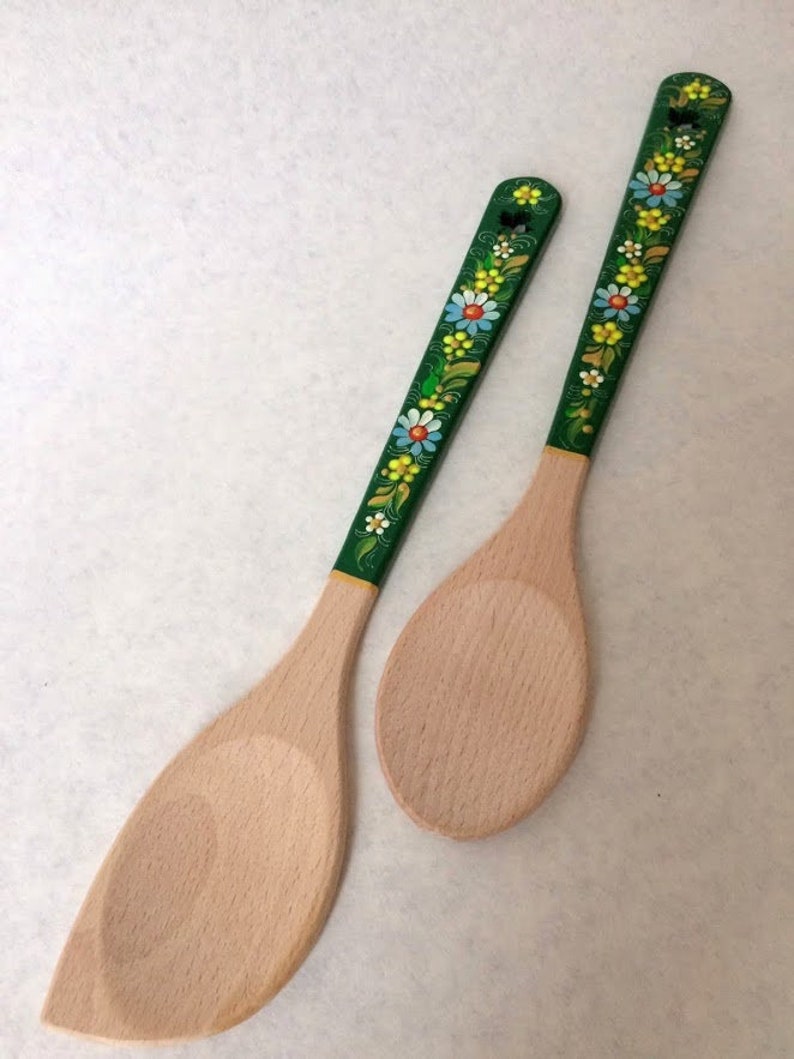 May include: Two wooden kitchen utensils: a spatula and a spoon. The handles are painted green and decorated with colorful floral patterns. The spatula is approximately 12 inches long, and the spoon is about 11 inches long.
