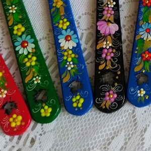 May include: A collection of hand-painted wooden spoons in various colors, including red, green, blue, and black. Each spoon is decorated with floral patterns in white, yellow, and pink, creating a vibrant and artistic display. The spoons are arranged on a white lace surface.