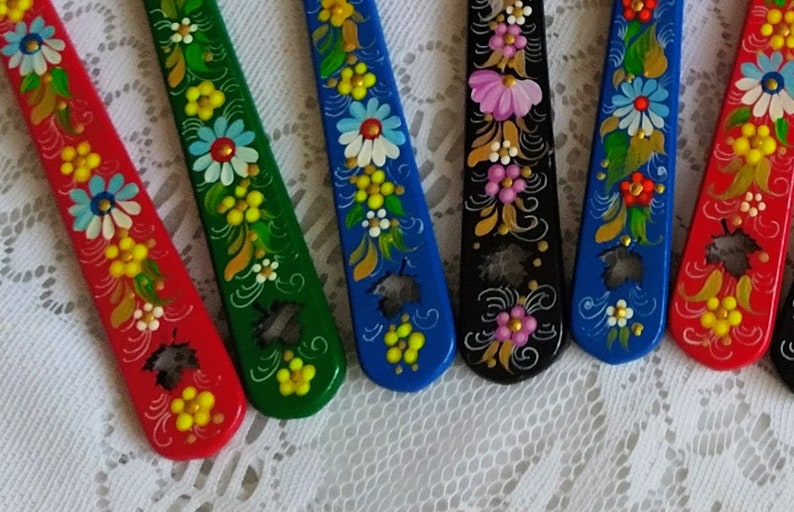 May include: A collection of hand-painted wooden spoons in various colors, including red, green, blue, and black. Each spoon is decorated with floral patterns in white, yellow, and pink, creating a vibrant and artistic display. The spoons are arranged on a white lace surface.