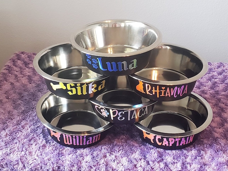Puede incluir: Cinco cuencos de acero inoxidable para mascotas apilados uno encima del otro. Los cuencos son negros con letras blancas. Los nombres en los cuencos son Luna, Sitka, Rhianna, Petaca y Captain.