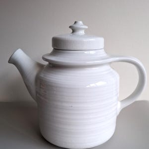 May include: A white ceramic teapot with a lid and handle. The teapot has a rounded body with horizontal ridges and a spout. The lid has a small knob on top. The teapot is on a grey surface.