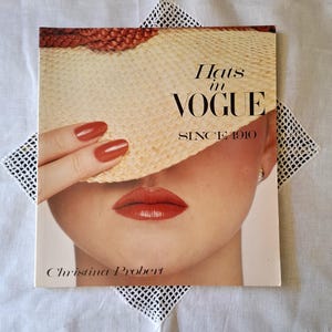 May include: A vintage magazine cover showing a woman with red lipstick and nails, holding a straw hat. The magazine title reads "Hats in VOGUE SINCE 1910" with the author's name, Christina Probert, at the bottom. The cover is set on a white, lace-trimmed surface.