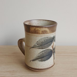May include: A brown and white ceramic mug with a leaf design. The mug has a handle and a brown rim.