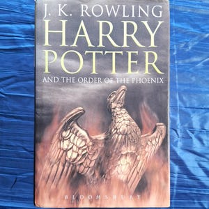 May include: A hardback book titled "Harry Potter and the Order of the Phoenix" by J.K. Rowling. The cover shows a phoenix with outstretched wings against a fiery backdrop. The publisher's name, Bloomsbury, is at the bottom.