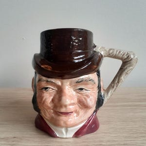 Puede incluir: Una taza de cerámica marrón con una cara y un sombrero marrón. La taza tiene un asa con forma de cuerda retorcida.