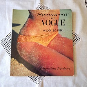 May include: A vintage book titled "Swimwear in VOGUE Since 1910" with a photograph of a person wearing an orange crochet bikini. The cover is dusted with sand, and the author's name, Christina Probert, is visible.