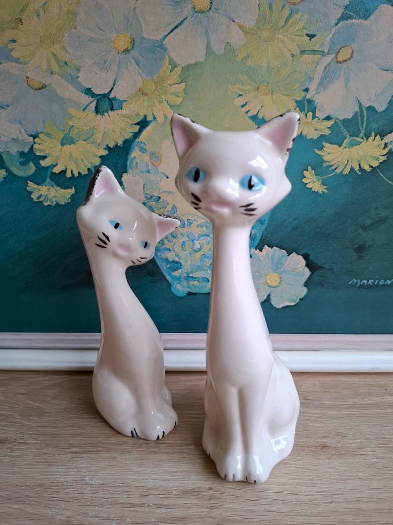 Pode incluir: Duas estatuetas de gato em cer&acirc;mica. Os gatos s&atilde;o brancos com detalhes pretos nas orelhas e bigodes. T&ecirc;m olhos azuis. Um gato est&aacute; sentado, o outro est&aacute; reclinado. O fundo apresenta uma pintura floral.