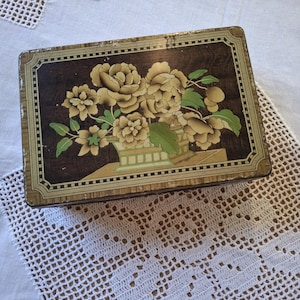 May include: A rectangular metal tin featuring a dark brown background with a central illustration of light yellow flowers in a green and white patterned vase. The lid has a decorative border with gold and black lines and shows signs of wear.