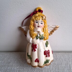 May include: A ceramic angel ornament with blonde hair, blue eyes, and gold-tipped wings. The angel wears a cream-colored dress adorned with red bows and green holly leaves. A burgundy ribbon is attached for hanging. The ornament has a gold trim.