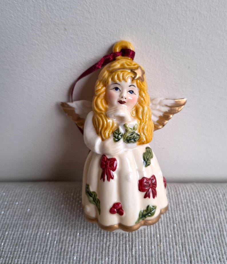 May include: A ceramic angel ornament with blonde hair, blue eyes, and gold-tipped wings. The angel wears a cream-colored dress adorned with red bows and green holly leaves. A burgundy ribbon is attached for hanging. The ornament has a gold trim.