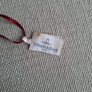 May include: A small, rectangular tag with the text "Villeroy & Boch 1748" printed in blue on a white background. The tag is attached to a thin, dark red ribbon. The background is a textured, light gray fabric.