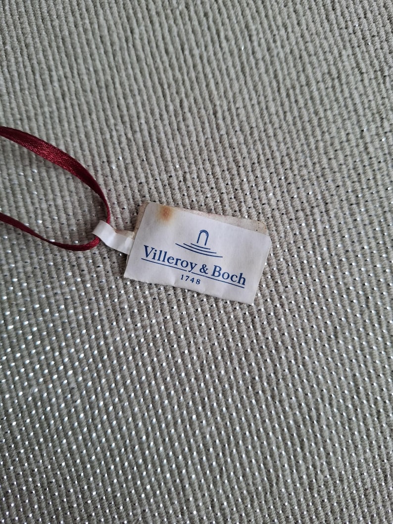 May include: A small, rectangular tag with the text "Villeroy & Boch 1748" printed in blue on a white background. The tag is attached to a thin, dark red ribbon. The background is a textured, light gray fabric.