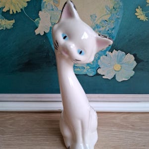 Pode incluir: Estatueta de gato em cer&acirc;mica branca com olhos azuis e detalhes em preto. O gato tem um pesco&ccedil;o longo e est&aacute; sentado. A estatueta est&aacute; sobre uma superf&iacute;cie de madeira, com uma pintura floral turquesa e branca ao fundo.