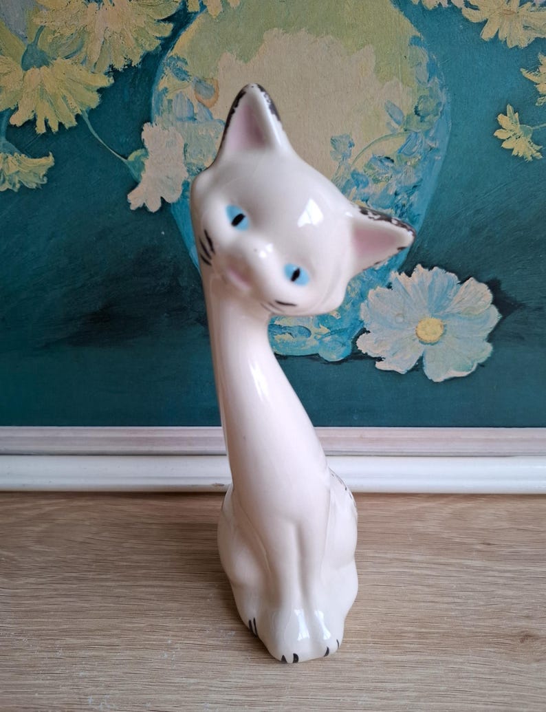 Pode incluir: Estatueta de gato em cer&acirc;mica branca com olhos azuis e detalhes em preto. O gato tem um pesco&ccedil;o longo e est&aacute; sentado. A estatueta est&aacute; sobre uma superf&iacute;cie de madeira, com uma pintura floral turquesa e branca ao fundo.