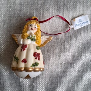 May include: A ceramic angel Christmas ornament with blonde hair, gold wings, and a cream-colored dress adorned with red bows and green holly. The ornament has a gold trim and a red ribbon for hanging. A tag from Villeroy & Boch is attached.