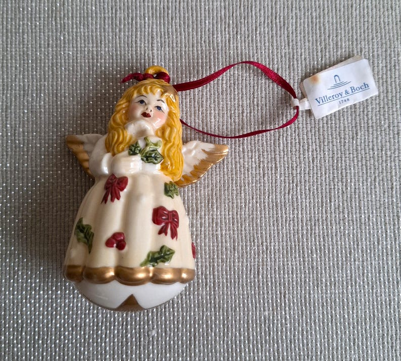 May include: A ceramic angel Christmas ornament with blonde hair, gold wings, and a cream-colored dress adorned with red bows and green holly. The ornament has a gold trim and a red ribbon for hanging. A tag from Villeroy & Boch is attached.