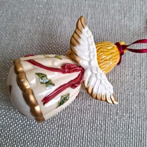 May include: A ceramic angel ornament with a cream-colored body, gold trim, and red ribbon accents. The angel has white wings with gold tips and a yellow head. A red ribbon is attached for hanging. Festive holiday decor.