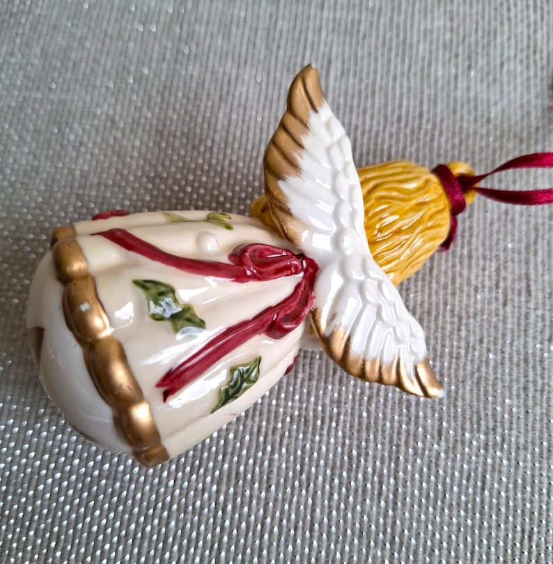 May include: A ceramic angel ornament with a cream-colored body, gold trim, and red ribbon accents. The angel has white wings with gold tips and a yellow head. A red ribbon is attached for hanging. Festive holiday decor.