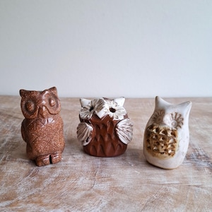 Könnte beinhalten: Drei Eulenfiguren aus Keramik in verschiedenen Braun- und Weißtönen stehen auf einer Holzoberfläche. Die Eulen haben strukturierte Oberflächen und detaillierte Merkmale, die eine rustikale Ästhetik zeigen.