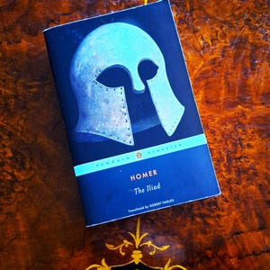 May include: A Penguin Classics edition of Homer's "The Iliad". The book features a dark blue cover with a light blue helmet illustration. The title and author's name are in white. The book is on a wooden surface.