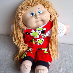 May include: A vintage doll with blonde yarn hair and blue eyes. The doll is dressed in a red jumpsuit with a white floral pattern and black detailing. The doll has a soft, round face and a gentle smile.