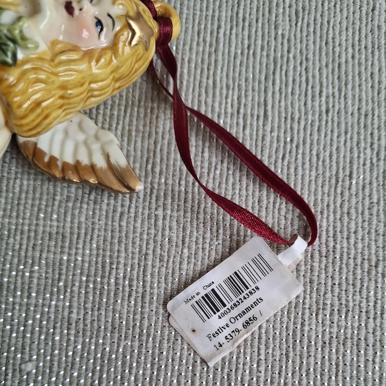 May include: A ceramic angel Christmas ornament with blonde hair, blue eyes, and gold accents. The angel has white and gold wings and is attached to a burgundy ribbon. A tag with a barcode and the words "Festive Ornaments" is attached to the ribbon.