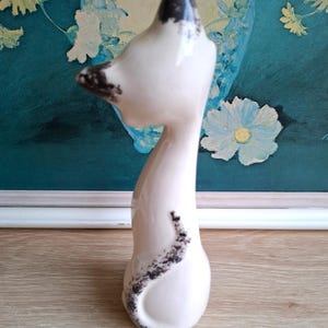 Pode incluir: Estatueta de gato em cer&acirc;mica creme com marcas pretas. O gato tem um pesco&ccedil;o longo e fino e uma cabe&ccedil;a pequena com orelhas pretas. A estatueta est&aacute; em p&eacute; e tem um acabamento liso e brilhante. &Eacute; um item decorativo.