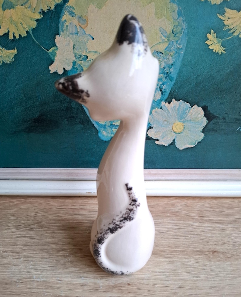 Pode incluir: Estatueta de gato em cer&acirc;mica creme com marcas pretas. O gato tem um pesco&ccedil;o longo e fino e uma cabe&ccedil;a pequena com orelhas pretas. A estatueta est&aacute; em p&eacute; e tem um acabamento liso e brilhante. &Eacute; um item decorativo.