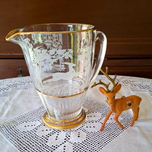 May include: A clear glass jug with a gold rim and base, featuring an etched forest scene with deer. A small, orange felt deer ornament with antlers and a floral detail is next to the jug. The jug is on a white lace tablecloth.