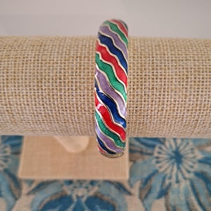 May include: A vibrant enamel bangle bracelet featuring a wavy striped design in red, green, purple, blue, and gold. The bracelet is displayed on a beige fabric surface.