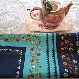 May include: A tea set with a floral design on a blue and navy tablecloth. The set includes a teapot, a glass bowl, a teacup, and a saucer. The tablecloth features a floral pattern with orange, yellow, and white flowers.