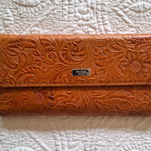 Könnte beinhalten: Eine braune Ledergeldbörse mit geprägtem Blumenmuster. Die Geldbörse hat eine rechteckige Form mit einem Klappverschluss. Der Markenname "Dechaz PARIS" ist auf einer kleinen silbernen Plakette zu sehen. Die Geldbörse liegt auf einer weißen, gesteppten Oberfläche.