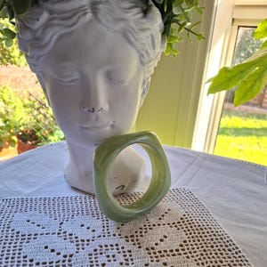 Może przedstawiać: Gładka bransoletka typu bangle z żywicy w kolorze szałwiowej zieleni o zaokrąglonym kwadratowym kształcie. Bransoletka ma błyszczące, półprzezroczyste wykończenie oraz gruby, masywny profil. Znajduje się przed białą rzeźbą popiersia w stylu klasycznym.
