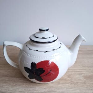 May include: White ceramic teapot with a black and red floral design. The teapot has a rounded body, a curved handle, and a spout. The lid is topped with a small knob and has black detailing. Suitable for serving tea.