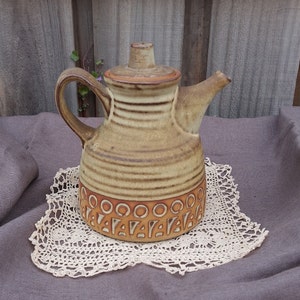 以下が含まれることがあります： 蓋と取っ手が付いたベージュのセラミックティーポット。水平線と幾何学模様のバンドで装飾されています。ティーポットは白いレースのドイリーの上に置かれています。背景は木製です。