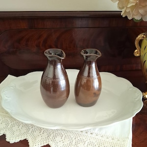 May include: Two brown ceramic vases with a dark rim and a textured surface. The vases are displayed on a white, scalloped-edge platter. The background features a dark wooden surface and a decorative vase with flowers.