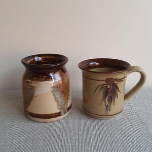 May include: Two ceramic pieces, a brown and beige mug with a floral design and a brown and beige jar with a brown rim.