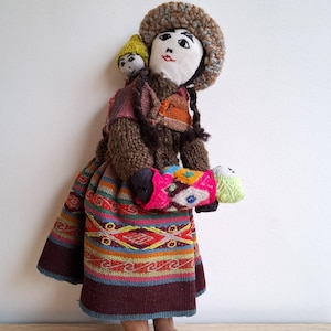 Può includere: Bambola fatta a mano con cappello e vestito marrone lavorati a maglia, che trasporta una bambola più piccola sulla schiena e un fagotto tra le braccia. La gonna della bambola presenta un colorato motivo geometrico con accenti rossi, blu e gialli. Il viso della bambola è dipinto in modo semplice.