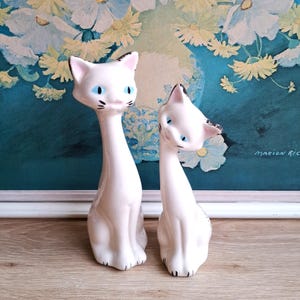 Pode incluir: Duas estatuetas de gato em cer&acirc;mica branca com detalhes pretos nas orelhas e patas, e olhos azuis. Um gato est&aacute; em p&eacute;, o outro est&aacute; inclinado. As estatuetas est&atilde;o sobre uma superf&iacute;cie de madeira, com uma pintura floral ao fundo.