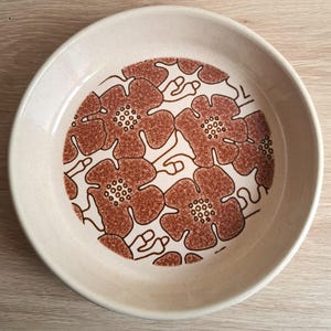 May include: A round, cream-coloured ceramic plate featuring a brown floral design. The flowers have a textured appearance and are arranged in a circular pattern. The plate's rim is a plain, neutral colour.