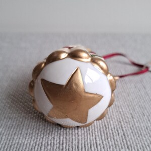 May include: A white and gold Christmas ornament with a gold star design. The ornament has a scalloped edge and a red ribbon for hanging. The star is painted gold.