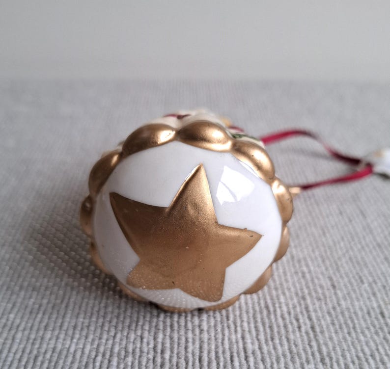 May include: A white and gold Christmas ornament with a gold star design. The ornament has a scalloped edge and a red ribbon for hanging. The star is painted gold.