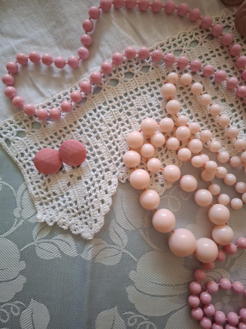 Może przedstawiać: Zestaw biżuterii składający się z długiego sznura jasnor&oacute;żowych koralik&oacute;w, kr&oacute;tszego wielowarstwowego naszyjnika z blado brzoskwiniowych koralik&oacute;w oraz pary okrągłych, fasetowanych kolczyk&oacute;w typu klips w kolorze przygaszonego r&oacute;żu.
