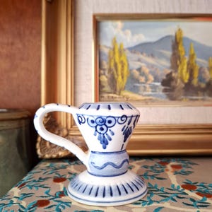 May include: A white ceramic candle holder with a handle, featuring hand-painted blue floral and geometric patterns. The design includes a flared base with vertical blue lines, a central band with a wavy line, and a top section with blue motifs.