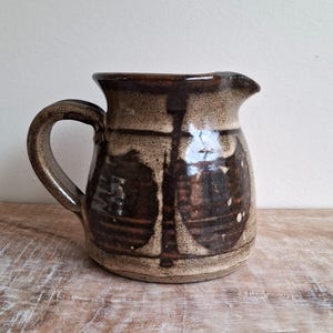 Può includere: Brocca in ceramica fatta a mano con smalto marrone scuro e beige. La brocca ha un corpo arrotondato, un manico curvo e un beccuccio. La superficie presenta motivi astratti marrone scuro e macchie bianche.